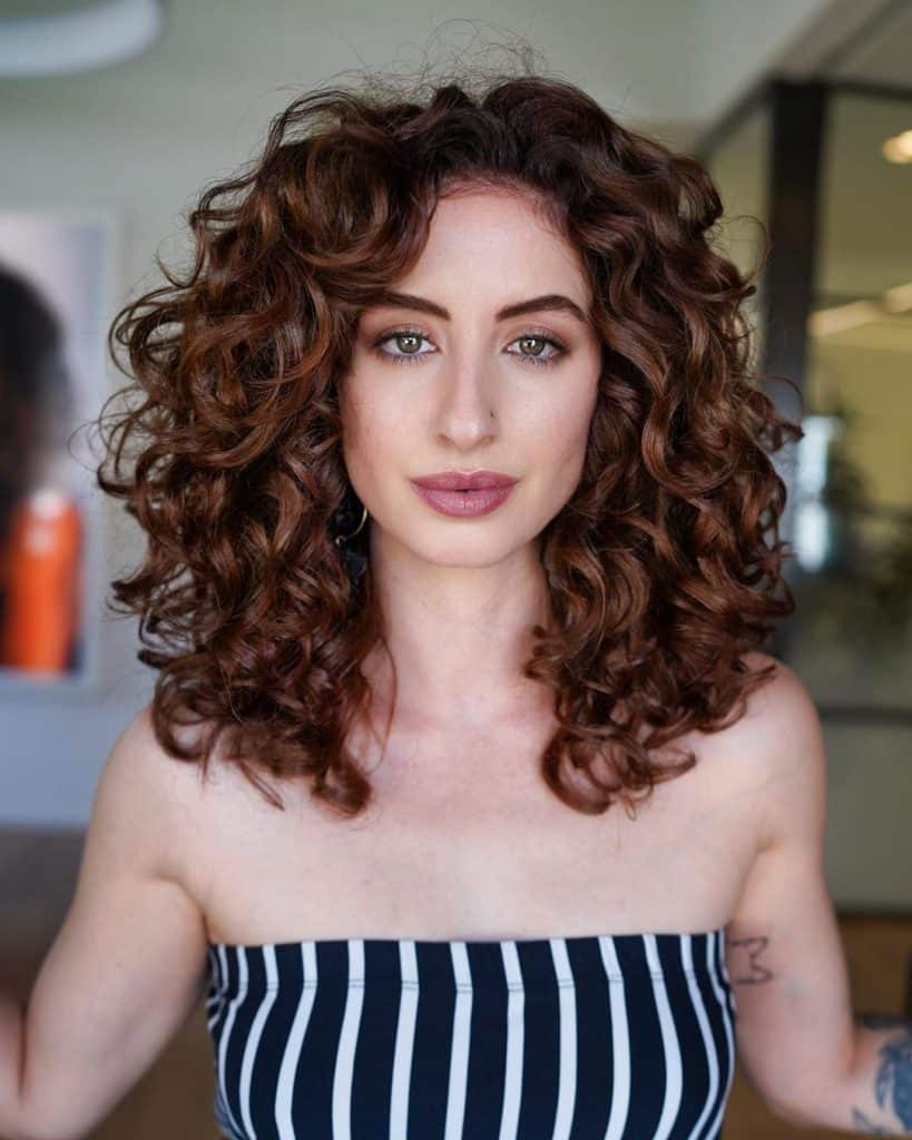 A woman with curly hair and tattoos, standing confidently with a serene expression on her face.
