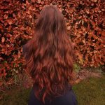 cheveux auburn A woman with long red hair standing in front of a bush in a serene garden setting.