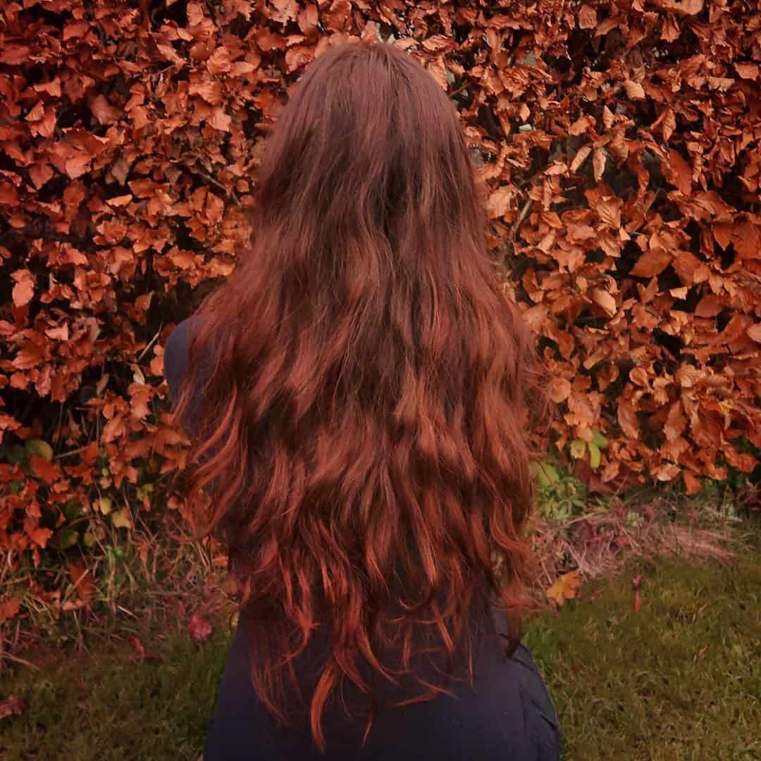 cheveux auburn A woman with long red hair standing in front of a bush in a serene garden setting.