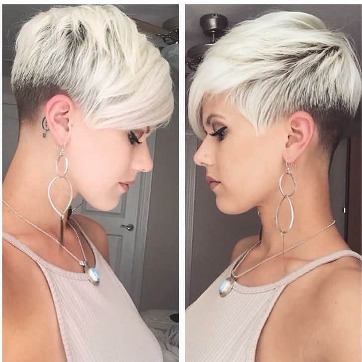 A profile view of a person with a stylish short blonde haircut, featuring an undercut and a long side-swept fringe, complemented by dangling earrings and a necklace.