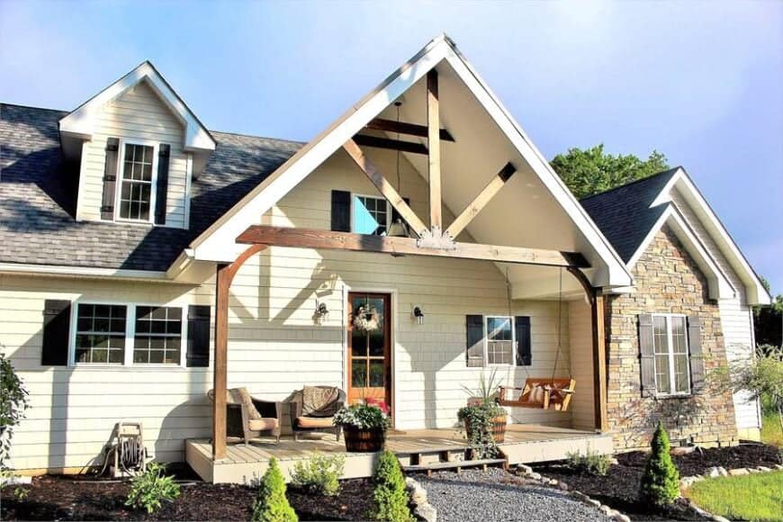 The covered front porch is filled with a swinging bench, two wicker round back chairs, and fresh potted plants.